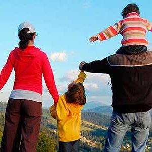 Family of four outside at the top of a grass hill looking down at a Forrest as being outside makes you happy.