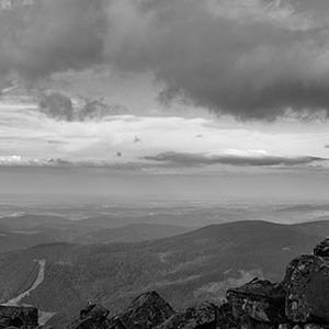 black and white photo of man sitting on cliff edge looking down into the valley.