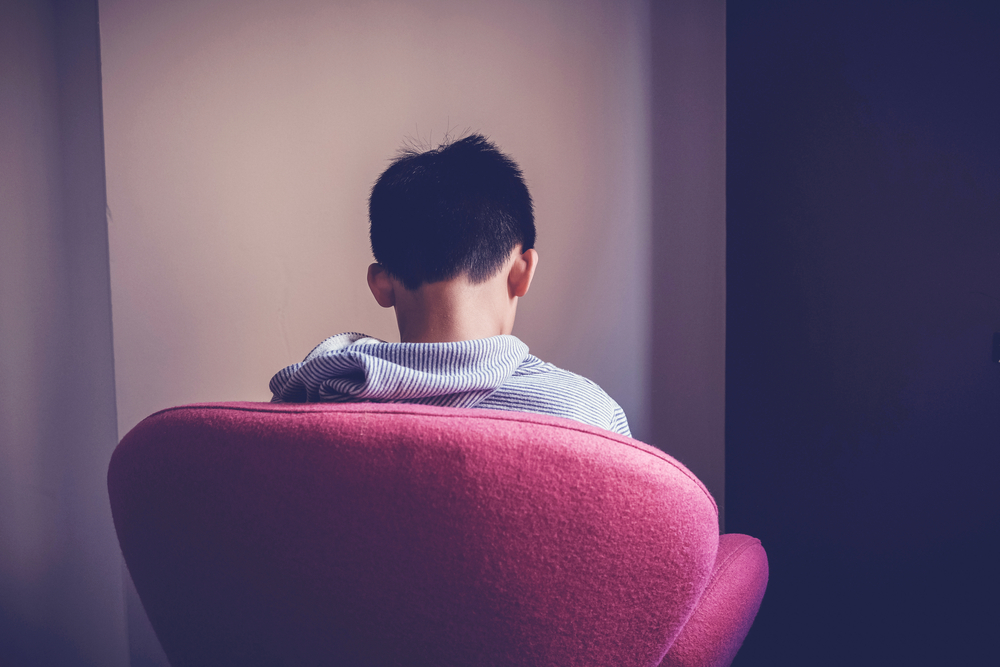 Sad preteen boy sitting alone in chair facing wall